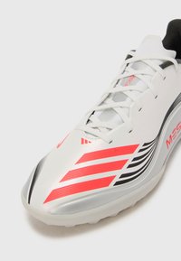 White athletic shoe with a sleek design, featuring red and black striped accents, synthetic material, and a textured sole for traction.