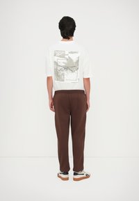 Camiseta blanca con estampado gráfico en la parte posterior, combinada con pantalones de chándal marrones. La camiseta presenta el logo de Reebok; los pantalones de chándal tienen cinturilla elástica.