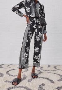Zwarte blouse met bloemenprint en wijde broek met witte details. Model draagt zwarte sandalen en houdt een kleine zwarte crossbody tas vast.