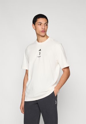 Camiseta de manga corta color crema hecha de algodón suave, con un logo negro de Adidas y la marca Juventus en el pecho, combinada con pantalones oscuros.
