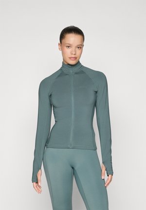 Kvinne med håret bundet i en hestehale iført en tettsittende turkis treningsjakke med glidelås og matchende tights, stående mot en ensfarget bakgrunn.