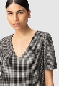 Młoda kobieta z włosami uczesanymi do tyłu, nosząca złote kolczyki w kształcie kół oraz czarno-białą bluzkę o wzorze houndstooth z dekoltem w serek i krótkim rękawem.
