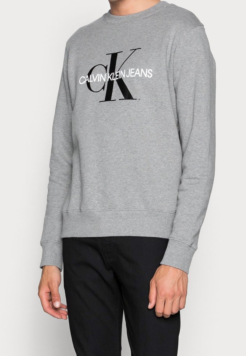 Personne portant un sweat-shirt gris avec le logo "Calvin Klein Jeans" en noir et blanc, associé à un pantalon noir.