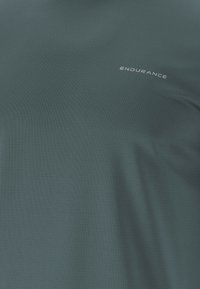 Tumman teal värinen urheilupaita, joka on valmistettu teksturoidusta kankaasta, sisältää sanan "KESTÄVYYS" valkoisena, sijoitettuna vasempaan yläkulmaan.