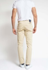 Hombre con pantalones beige, zapatillas blancas y una camisa blanca de manga corta de pie de espaldas contra un fondo blanco liso.