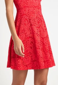 Röd spetsklänning med blommiga och paisleymönster, åtsittande vid midjan, utsvängande i en kort A-linjeformad kjol, bärs av en stående person.