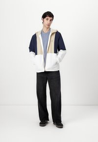 Kleurblokken jack in marineblauw, beige en wit; voorzien van een capuchon, ritssluiting aan de voorkant en een lichte stof. Gecombineerd met zwarte rechte jeans.
