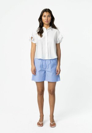 Žena stojaca v bielej blúzke s krátkym rukávom na zapínanie na gombíky, modro-bielych pruhovaných šortkách a bielych sandáloch na jednoduchom pozadí.