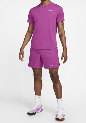 Hombre de pie con camiseta deportiva y pantalones cortos morados de Nike, calcetines blancos y zapatillas Nike blancas y moradas sobre fondo blanco.