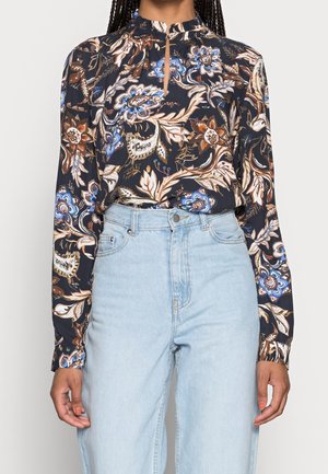 Kobieta w ciemnej, kwiecistej bluzce z długim rękawem, wpuszczonej w jasnoniebieskie, wysokowyższe dżinsy, z widocznymi splecionymi warkoczami.