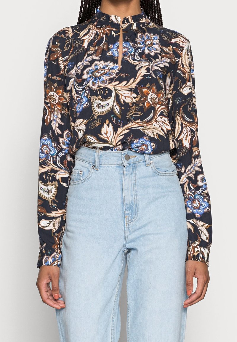 Femme portant un chemisier à manches longues à fleurs sombres, rentré dans un jean taille haute bleu clair, avec des cheveux tressés visibles.