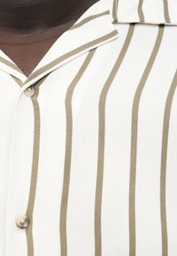 Close-up van een donkerhuidig persoon dat een wit shirt draagt met verticale beige strepen en een kraag met knopen.