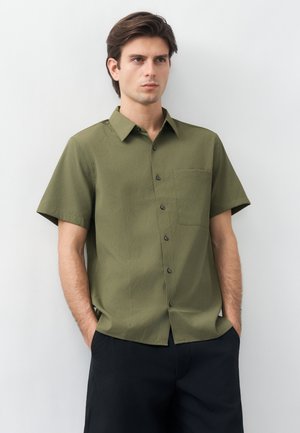 Νεαρός άνδρας με καστανά μαλλιά, φορώντας πουκάμισο σε ελιά πράσινο με κοντά μανίκια και μαύρο παντελόνι, στέκεται μπροστά σε έναν λευκό τοίχο.