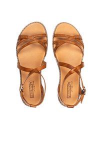 Sandalias de cuero marrón con correas entrelazadas, correa ajustable en el tobillo y acentos de tachuelas metálicas a lo largo del borde de la suela. Forro interior suave.