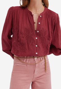 Chemisier bordeaux avec broderie présentant des motifs floraux, associé à un jean rose clair et une ceinture en chaîne dorée. Tissu texturé.