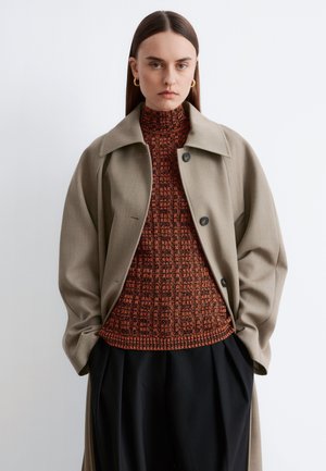 Femme aux cheveux longs et raides portant des boucles d'oreilles créoles en or, un manteau beige par-dessus un pull à col roulé rouge à motifs, et un pantalon noir.