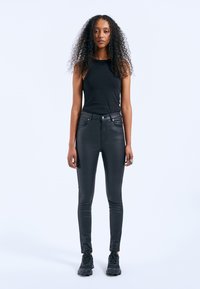 Jeans neri aderenti con una trama liscia, vita alta e cinque tasche. Modello abbinato a un top senza maniche nero e sneakers nere.
