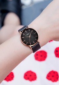 Reloj de pulsera negro con una caja redonda en oro rosa y una correa de malla negra. Presenta un dial minimalista con marcadores de hora y agujas en oro rosa.
