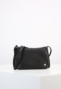 Černá kožená crossbody taška se štíhlým nastavitelním popruhem a malým stříbrným logem vpředu, umístěná na bílé podstavě proti jednoduchému pozadí.