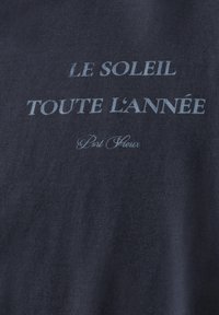 Tmavě modrá tkanina s tištěným textem. Text zní: "LE SOLEIL TOUTE L'ANNEE" v tučných a kurzivních písmenech, s "Port Fleur" v menších písmenech.