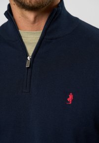 Navyblauer Reißverschluss-Pullover mit geripptem Kragen, der ein kleines rotes Logo auf der Brust aufweist. Weiches Material, körperbetontes Design und glatte Textur.