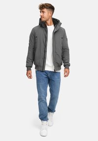 Chaqueta bomber gris con capucha, cierre de cremallera y bolsillos laterales. Llevada sobre una camisa blanca y combinada con jeans azules y zapatillas blancas.