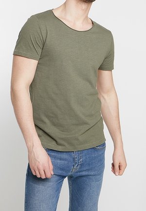 Mand iført en olivengrøn kortærmet t-shirt og blå jeans, stående med armene afslappet mod en ensfarvet lys baggrund.
