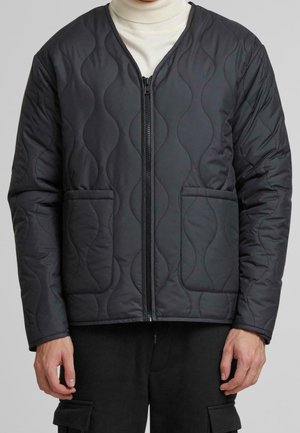 Homme portant une veste matelassée noire zippée avec deux poches avant et un col roulé blanc en dessous.