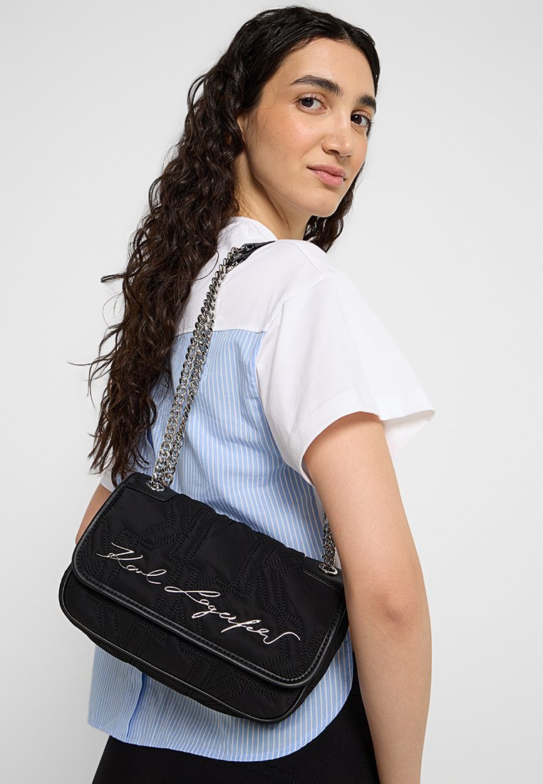 Sac à main noir matelassé avec texte brodé, chaîne en bandoulière, forme rectangulaire et texture lisse. Porté sur l'épaule.
