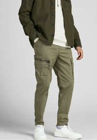 Man som bär olivgröna cargobyxor med dragkedje- och fläpplommar, vita sneakers, krämfärgad hoodie och mörk olivgrön knäppjacka.