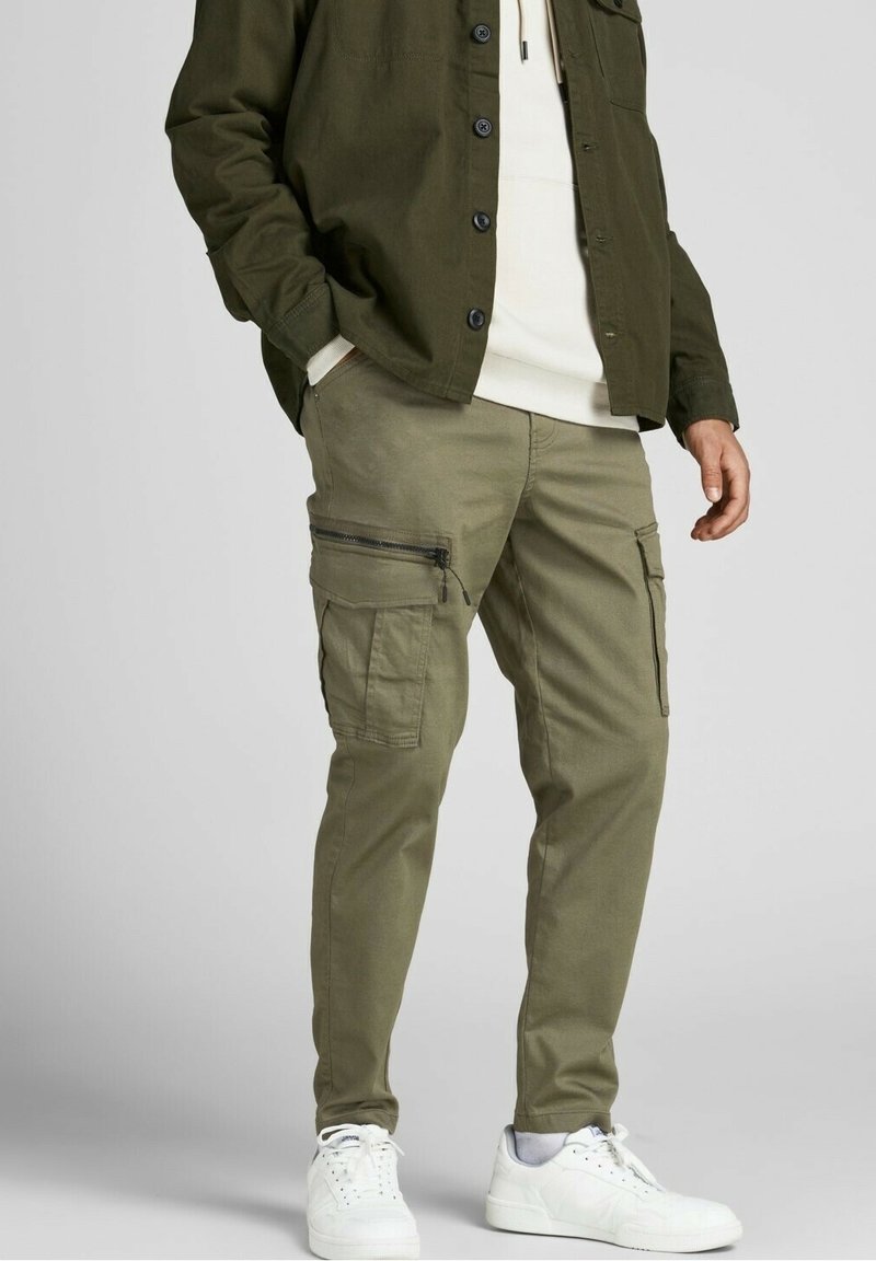 Man som bär olivgröna cargobyxor med dragkedje- och fläpplommar, vita sneakers, krämfärgad hoodie och mörk olivgrön knäppjacka.