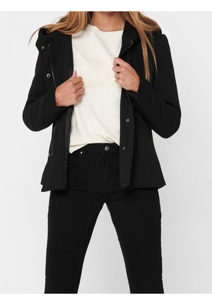 Femme portant une veste noire par-dessus un pull blanc et un pantalon noir, tenant la veste ouverte avec les deux mains.