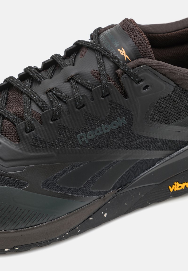 Scarpa sportiva nera con parte superiore testurizzata, chiusura con lacci, marchio Reebok e suola Vibram con elemento di design maculato.