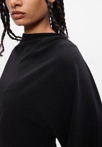 Top negro con un cuello alto y un diseño drapeado. Tela suave, corte holgado y detalles de costura sutiles a lo largo del hombro y el costado.