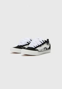 Schwarze und graue Sneakers mit einem Muster, weißen Schnürsenkeln, einem flachen Design, Gummisohle und kontrastierenden weißen Streifen.