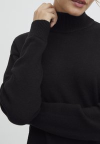 Juodas turtleneck megztinis, pagamintas iš minkštos mezgimo medžiagos, turintis guluolus rankogalius ir apykaklę, demonstruojantis laisvą siluetą ir minimalistinį dizainą.