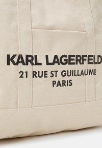 Borsa a sacca in tela color beige con testo stampato in nero: "KARL LAGERFELD 21 RUE ST GUILLAUME PARIGI." Presenta manici robusti e cuciture rinforzate.