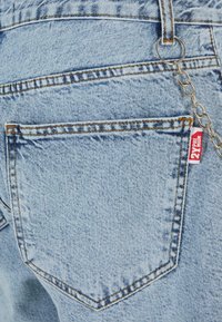 Gai zila denim bikses ar aizmugurējo kabatu, kas rotāta ar oranžām šuvēm, un ķēdi ar sarkano birku, kas piesaistīta jostasvietai.