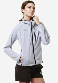 Giacca zip grigia chiara con cappuccio, caratterizzata da una texture a griglia, zip nera e taschino sul petto. Abbinata a pantaloni corti neri.