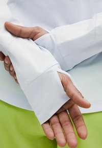White long-sleeve shirt with thumbholes in the cuffs, smooth texture, and fitted design. Green shorts visible in the lower frame.