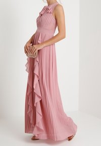 Roze plissé maxi jurk met ruches, zonder mouwen en een getailleerde taille. Gestyled met een metallic clutch en sandalen.