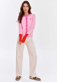 Różowy i czerwony kardigan z zapięciem na guziki, z detalem w kształcie serca i długimi czerwonymi mankietami. W zestawieniu z jasnymi spodniami i butami w panterkę. Gładka faktura.