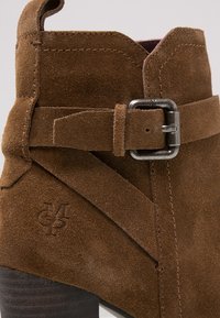 Brune ruskinds ankelstøvle med et spidst design. Har et metalspænde som accent og præget logo på siden. Mørkt træhæl.