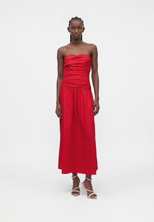 Γυναίκα που στέκεται με λευκές ψηλοτάκουνες σανδάλια με λουράκια και φορά ένα κόκκινο midi φόρεμα χωρίς τιράντες και με ρεβέρ, στο φόντο ενός μονόχρωμου σκηνικού.