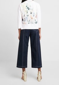 Světle růžový svetr s květinovým potiskem na zadní straně, spárovaný s širokými námořnickými culottes. Model má na sobě zlaté sandály s blokovým podpatkem.