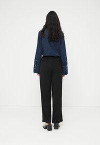 Σκούρο τζιν πουκάμισο με μακριά μανίκια, μαύρο παντελόνι με φαρδιά μπότα, και μαύρα παπούτσια με χοντρό τακούνι. Τα μαλλιά είναι μακριά και κυματιστά. Ουδέτερο φόντο.