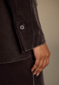 Ciemna aksamitna marynarka z guzikami na mankiecie, charakteryzująca się gładką fakturą i subtelnym połyskiem. Zbliżenie przedstawia dłoń spoczywającą na tkaninie.