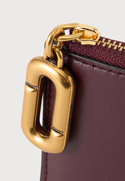 Gros plan d’un fermoir en métal doré fixé à une pochette zippée en cuir violet foncé avec un ruban de fermeture éclair en tissu violet assorti.