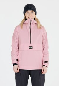 Rosa wasserfeste Jacke mit einer Fronttasche, schwarzen Reißverschlüssen und schwarzen Bündchen. Kombiniert mit einer dunklen Strickmütze mit Logo.