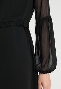 Schwarzes Kleid mit durchsichtigen, puffärmeligen langen Ärmeln mit gerafften Bündchen und einem gerafften Taillenbund, gefertigt aus leichtem Stoff mit strukturierter Oberflächenbeschaffenheit.